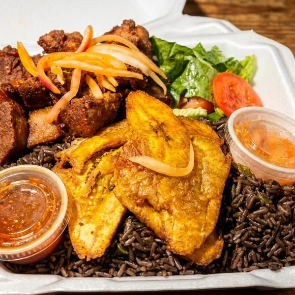 Haitian griot with plantains, black rice, salad, and two sauces in a takeout container.